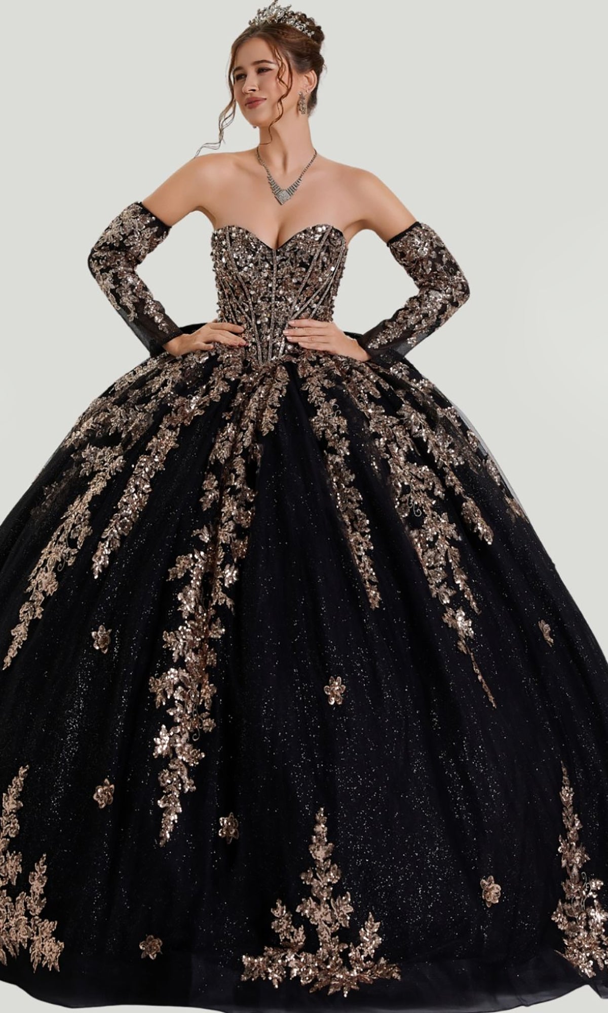 A woman in the Quinceanera Dress 26402 by Stella Couture, a stunning black strapless ball gown adorned with gold floral embellishments, featuring a beaded corset bodice and a glitter tulle skirt, accessorized with matching gloves and a necklace, poses gracefully with her hands on her waist.